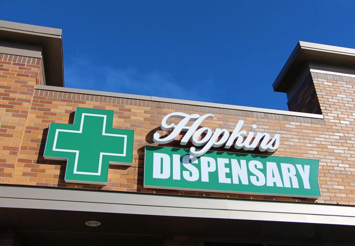 Hopkins Dispensary storefront — green cross and Hopkins Dispensary sign on brick against a blue sky.